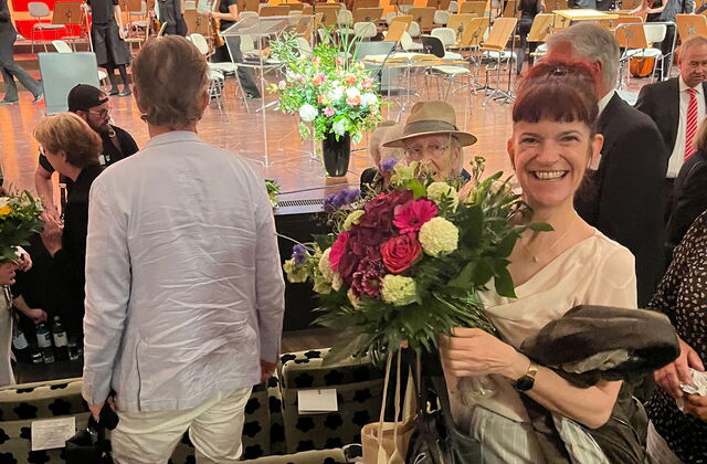 Eine Frau mit einem großen Blumenstrauß in heller Bluse sowie Hose. Sie steht in einer Sitzreihe des Nikolaisaals uf der Bühne sind noch Musiker.