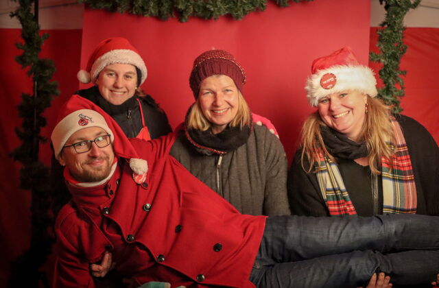 Drei Frauen lächeln, ein Mann liegt über deren queer auf ihren Beinen. Sie haben rote Weihnachmützen auf. Der Mann trägt eine Brille. Sie posieren vor einem roten Hintergrund.
