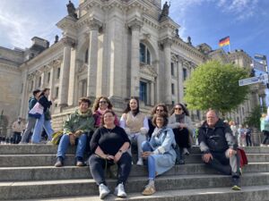 Gruppenbild: 7 Frauen und ein Mann sitzen auf Stufen vor dem Reichstag in Berlin. Alle lächeln fröhlich.