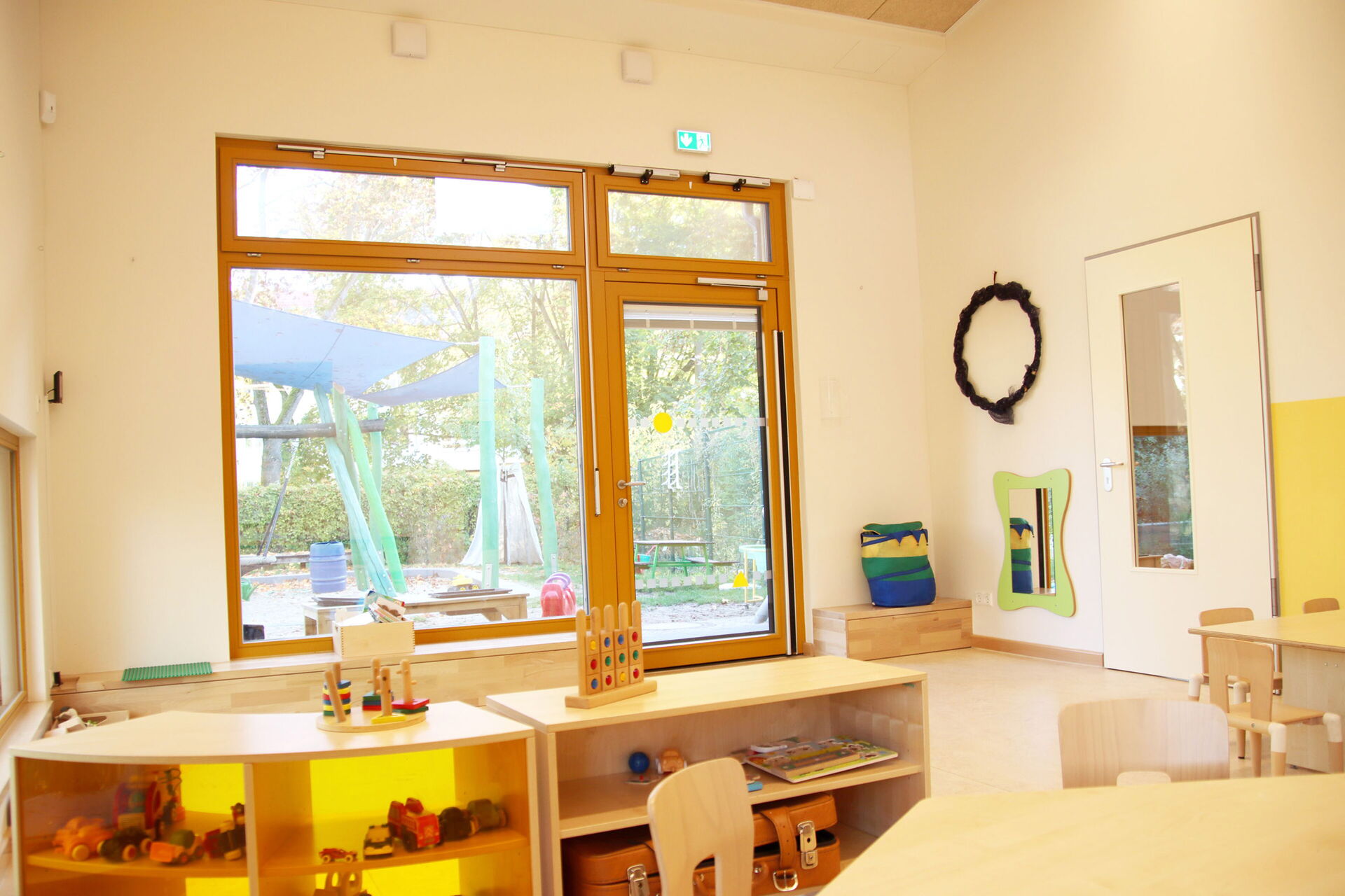 Spielraum. Großes Fenster, Terrassentür, die zu einem kleinen Spielplatz führt. Im Raum, zwei kleine Regale mit Spielzeug, zwei kleine Tische mit Stühlen.