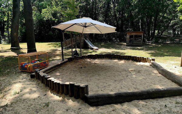Sandkasten mit Sonnenschirm. Im Hintergrund Bäume.