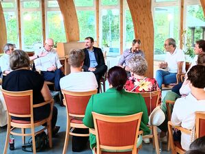 16 Menschen sitzen auf Stühlen locker verteilt kreis ähnlich, so das man sich ansehen und gut austauschen kann. Es werden Gespräche geführt.