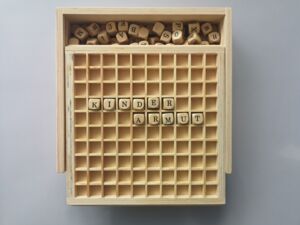 solide Holzbox mit Schiebedeckel beinhaltet Buchstabenwürfel, die in Wörter und Kreuzwortgebilde gelegt werden. Das Wort Kinderarmut ist über zwei Reihen gelegt.
