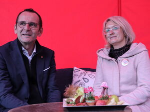Ein Mann und eine Frau sitzen auf einem Sofa vor einer roten Wand vor ihnen auf einem runden Tisch ein herbstlich dekoriertes Gesteck.