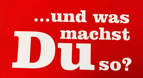 Rote Schrifttafel mit weißer großer Schrift. Der Werbetext der AWO Ehrenamtsagentur fragt: ... und was machst Du so?