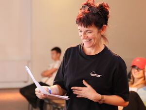 Frau mit roten Haaren, Brille mit scharzem Rahmen hat sie auf den Kopf geschoben, sie trägt ein schwarzes T-Shirt, eine Armbanduhr am linken Kangelenk, sie lächelt. Sie hält Notizen in der Hand. Im Hintergrund sitzen Jugendliche.