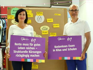 Eine Frau und ein Mann stehen vor einer Flipchart-Wand und halten jeweils ein lila Schild in den Händen. Beim Mann die Forderung: Kostenloses Essen an Kitas und Schulen.