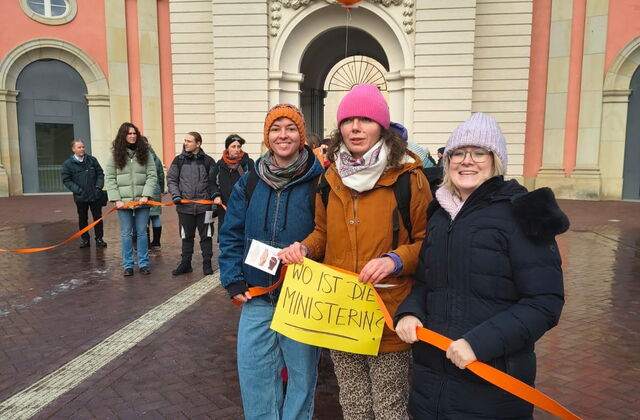 Frauen halten ein orangenes Band. 3 Frauen im Vordergrund mit orangenem Luftballon und einem Schild in den Händen. Text: Wo ist die Ministerin?