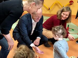 Ein kleines Mädchen tapst auf einen alten Mann mit Glatze im dunklen Anzug und weißem Hemd darunter zu. Der Mann streckt ihr die Hand entgegen. Eine Frau beugt sich vor. Eine Frau mit Kind auf dem Schoß sitzt auf dem Fußboden. Eine weitere Frau hockt nebe