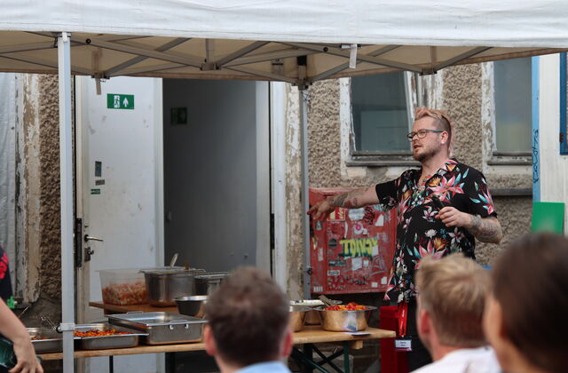 Mann mit dunklem Hawaii Hemd zeigt auf das fertige Essen auf Tischen unter einem Pavillon.