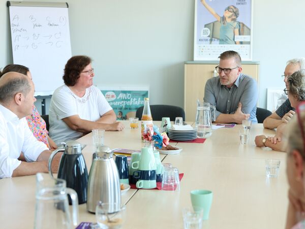 7 Personen an einem großen Tisch. Auf dem Tisch, Teller, Tassen, Gläser und Thermoskannen. An der Stirnseite ein Mann mit Brille und grauem Hemd, 2 Frauen und der Bundeskanzler sitzen links am Tisch.