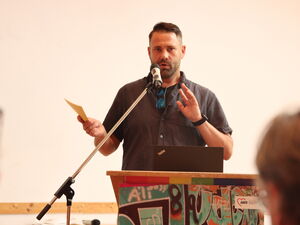 Mann am Rednerpult. Er hat Karteikarten / Notizen in der rechten Hand und sprich in ein Mikrofon.