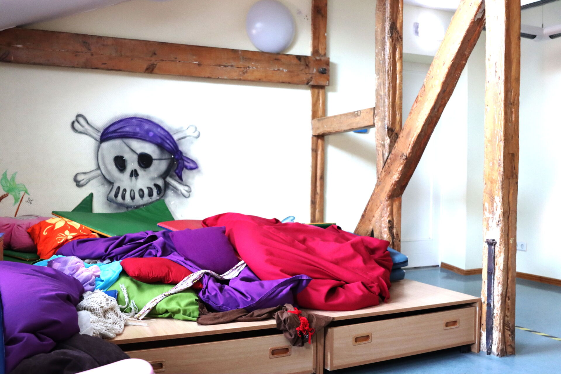 Holzbalken stützen den Raum. Eine Lümmelecke mit vielen farbigen Kissen und Decken, an der Wand dort ein Piraten-Totenkopf mit lila Kopftuch.