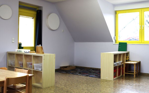 Spielzimmer. In der Ecke liegt ein Straßenteppich. Zwei kleine Regale stehen bei den Fenstern. Ein kleiner Tisch mit kleinen Stühlen.