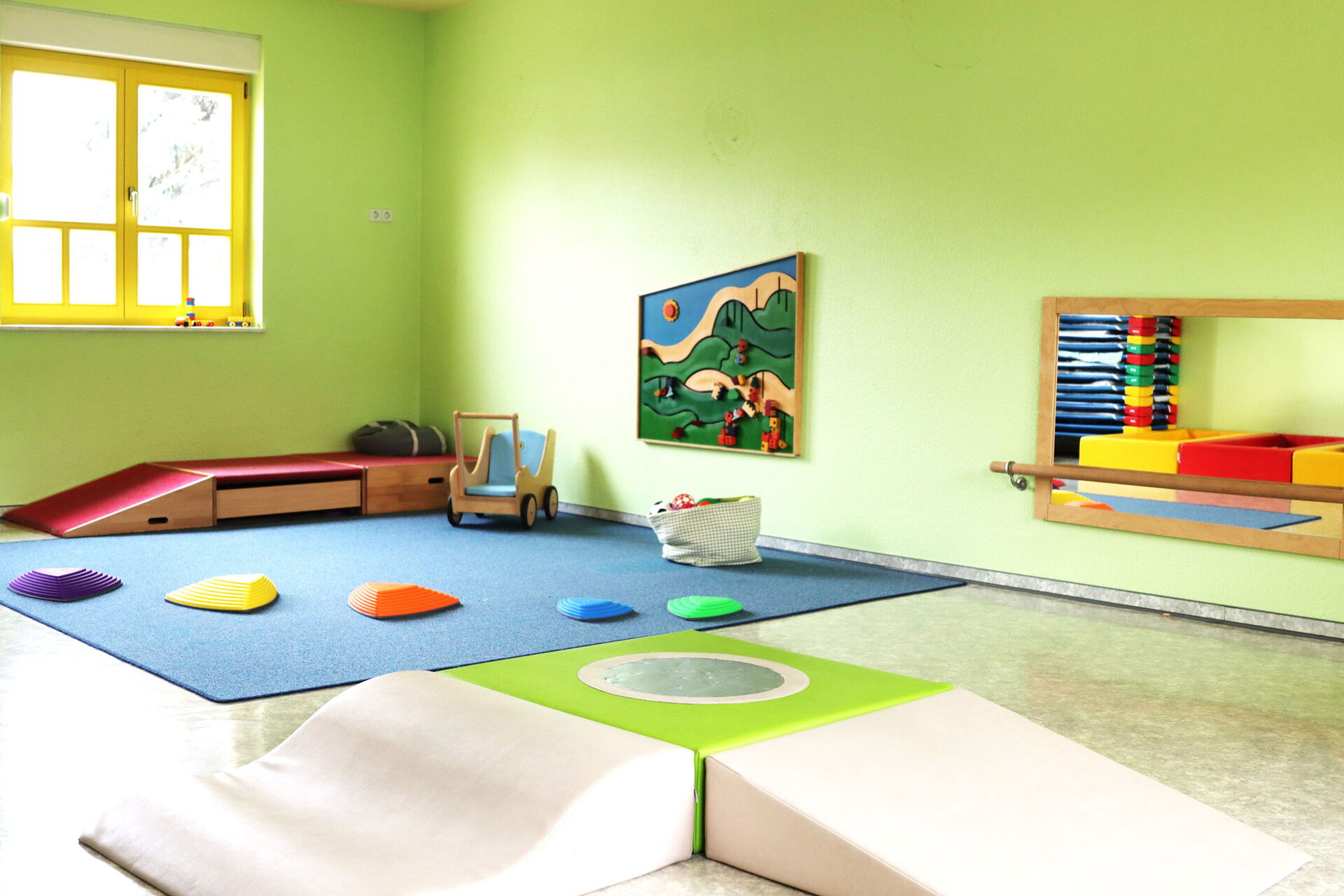 Spielzimmer. Großer Raum mit grünen Wänden. Auf einem blauen Teppich der in der Ecke liegt sind farbige Balanciersteine Für Kinder (lila, gelb, orange, blau, grün) in unterschiedlichen Größen An der Wand ein Spiegel.