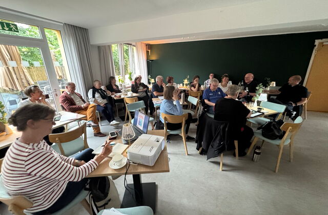 Sitzung: Größerer Raum, Menschen sitzen locker an Tischen.  Vor im Bild eine Frau an einem Tisch mit Laptop und Beamer.