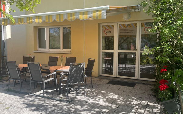 Terrasse mit Stühlen und Tischen. Eine Markise spendet Schatten, die Blumenkästen sind mit Geranien bepflanzt.