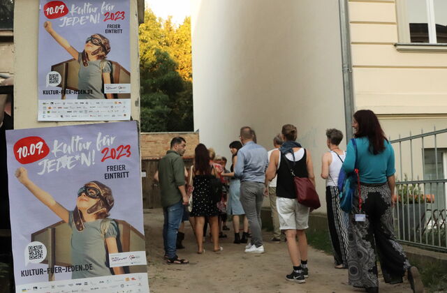 Draußen. Häuser. Große Plakate von Kultur für Jeden an Hauswand. Eine kleine Gruppe Mensch in sommerlicher Kleidung.