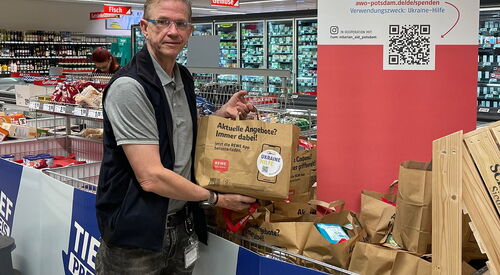 Ein Mann in einem Rewe Markt mit einer gefüllten Papiertüte in den Händen. Es stehen schon mehrere solcher Tüten in einem dafür vorgesehenen Verkaufsständer.