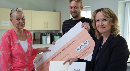 Drei Personen in einem Raum. Blonde Frau und Mann mit Vollbart halten einen großen Scheck über 64.385,90 Euro in den Händen. Frau mit braunen gewellten schulterlangen Haaren und dunklem Blazer hält eine Urkunde in die Kamera.