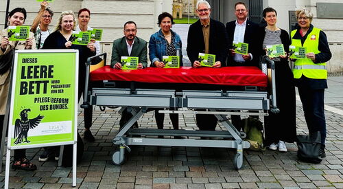 vor dem Fortuna Portal. Ein leeres Pflegebett, ein großes grünes Schild und 10 Menschen mit grünen Karten, mit Aufschrift.
