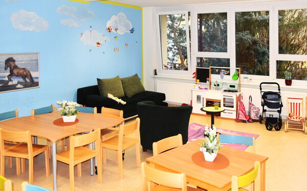 Hell, freundlich eingerichteter Raum, Spielzimmer. Die Wände sind hellblau gestrichen und mit einer Wandmalerei von Wolken und Vögeln verziert. Es gibt zwei Tische mit Stühlen.Ein schwarzes Sofa und Sessel stehen in der Ecke am Fenster.