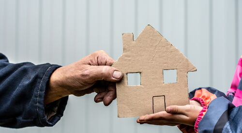 Zwei gegenüberstehende Personen halten jeweils mit einer Hand gemeinsam ein kleines Haus aus Pappe, im Hintergrund eine helle Wand.