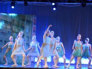 Schauspielerinnen auf der Bühne tanzen eine Choreographie. Sie haben hellblaue Kleider an und tragen Kronen.