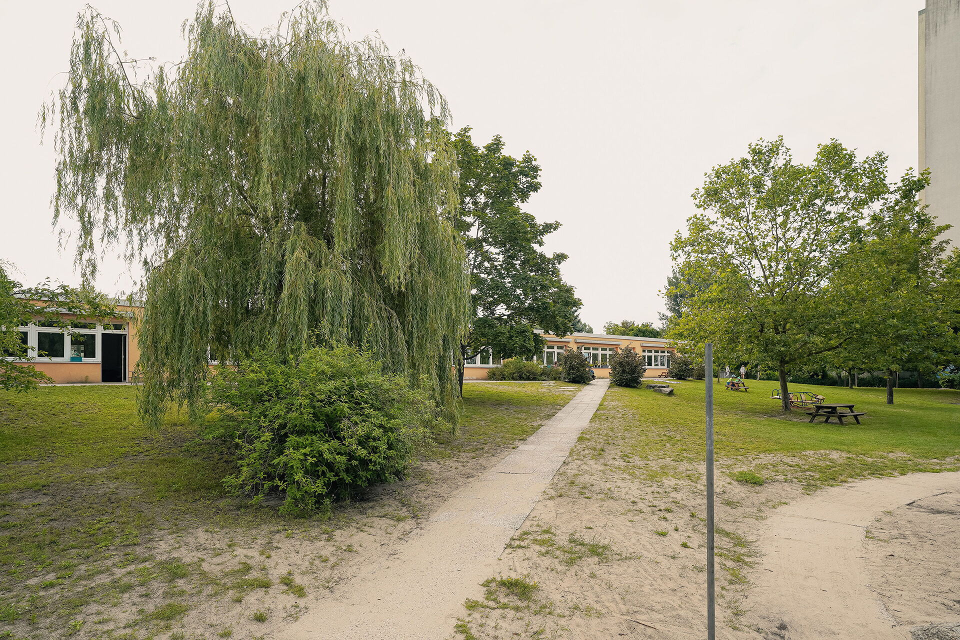 Draußen, ein Weg führt zu einem flachen langen Gebäude, Bäume und Sträucher und Wiese.