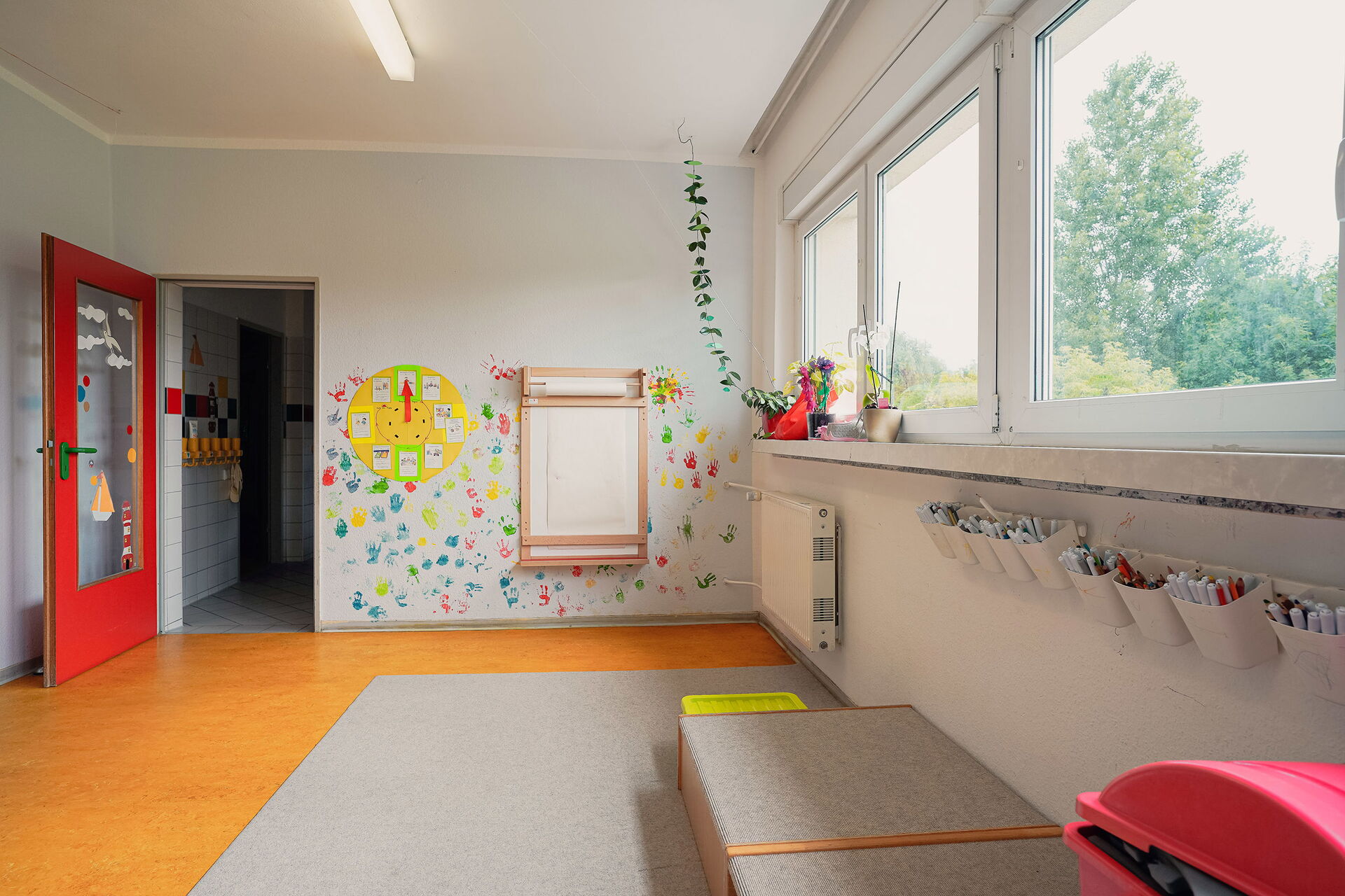 Raum, rote Tür steht offen. Im Raum ist ein hellgrauer Teppich. An der Wand neben der roten Tür ist eine gebastelte Uhr, und an der Wand sind farbige Handabdrücke der Kinder in blau, gelb, rot und grün.