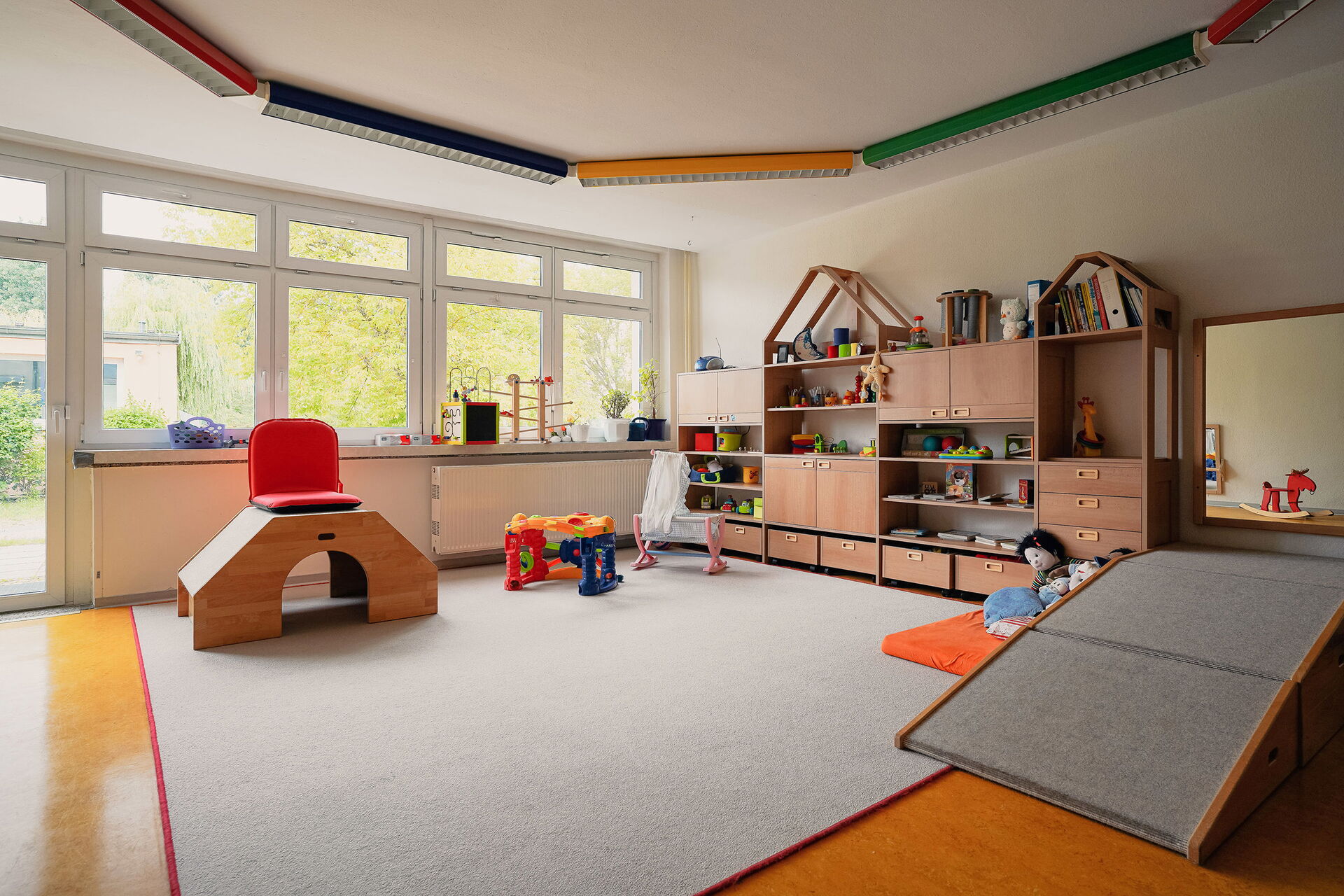 Spielzimmer. Regal mit Spielsachen an der Wand. Ein hellgrauer Teppich liegt im Zentrum des Raums. Puppenbettchen und Puppen.