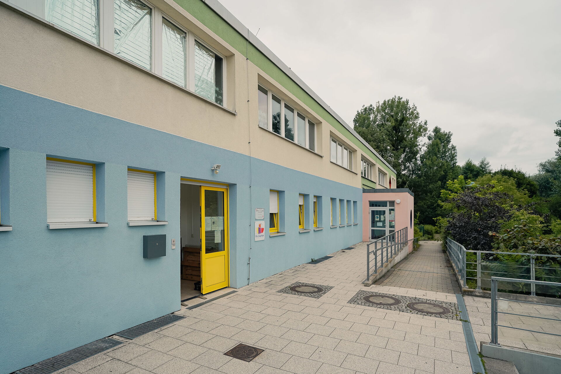 Außen. Gelbe Tür zum Gebäude steht offen. Eine Rampe führt zum Weg hinab. Das Gebäude hat zwei Stockwerke die Fassade ist unten blau, darüber beige und darüber grün.