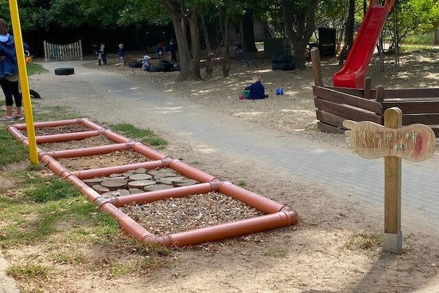 Spielplatz mit roter Rutsche im Vordergrund ein Barfußpfad. Kleine Kinder spielen auf dem Spielplatz.
