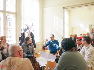 Großer Seminarraum. Personen sitzen im Kreis. Ein leeres Flipchart-Papier liegt in der Mitte der Personen auf Stühlen / Tischersatz.