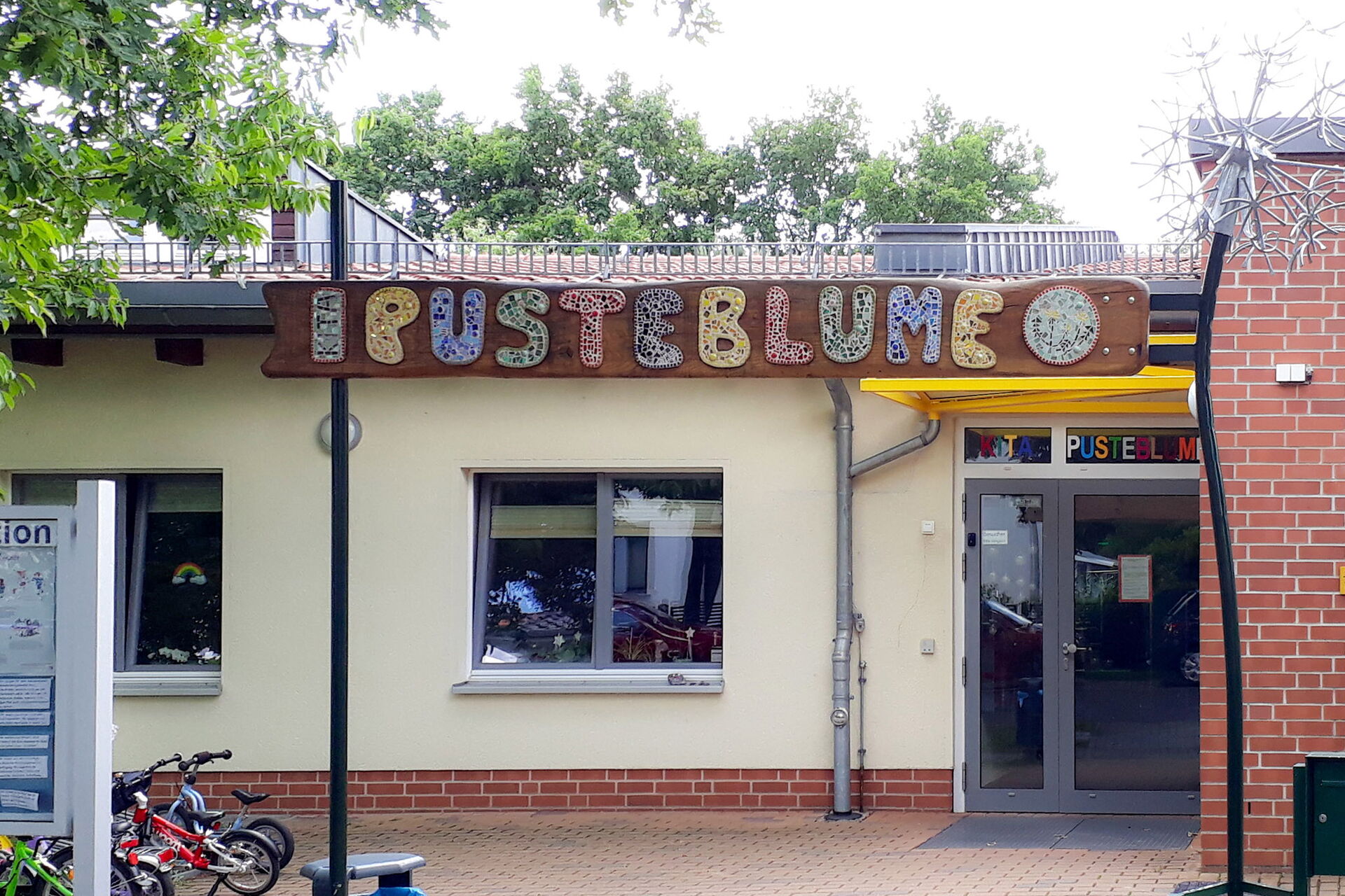 Eingang einer Kita. In großen farbigen Buchstaben über dem Eingang steht das Wort: PUSTEBLUME