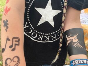 Mensch mit schwarzem T-Shirt mit großes Stern. Ausgestreckte Arme. Auf den Armen Airbrushtattoos: ein Herz, Noten, eine Fee und Anderes ums Handgelenk ist das blaue Festival-Bändchen.