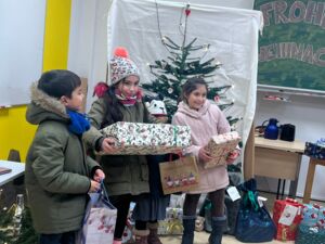 Drei Kinder in Winterkleidung  im Klassenraum vor dem beleuchteten und geschmückten Tannenbaum. Ein Junge und zwei Mädchen haben Geschenke und Geschenktüten in den Händen und freuen sich darüber.