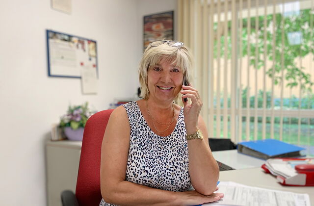 Frau an einem Streibtisch sitzend. Mit Telefon am Ohr. Frau ist blond, Brille auf den Kopf geschoben.