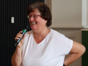 Frau in weißem T-Shirt und Brille spricht fröhlich in ein Mikro.