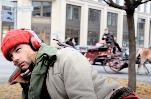 Draußen in einer Stadt. Ein Mann mit roter Mütze und roten Kopfhörern, graue Jacke. Im Hintergrund eine Pferdekutsche.