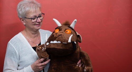 Altere Frau, mit grauen kurzen Haaren, Brille, Ohrstecker und Kette mit heller Strickjacke lächelt ihre zwei Grüffelos an. Ein kleines und ein großes braunes Kuscheltiermonster mit großen Zähnen, Hörnern und orangenen Augen.