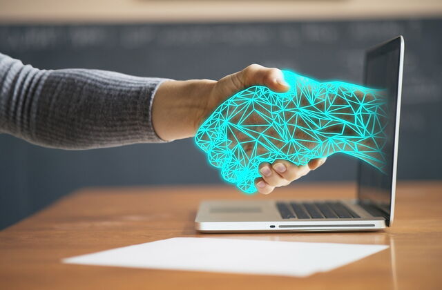 Laptop auf einem Tisch, aus dem Bildschirm ragt eine leuchtend blaue Cyber-Hand heraus und schüttelt eine menschliche Hand, die von links entgegen gestreckt wird.