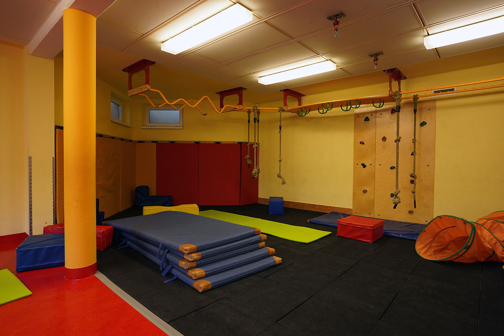 Turnhalle. 5 blaue Matten sind übereinander gestapelt. Eine Kletterwand mit Tritten. Kletterparcours an der Decke befestigt mit Stangen und unterschiedlichen Griffen.