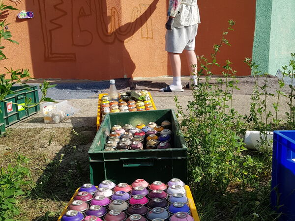 Mehrere bunte, bemalte Holzleisten liegen auf einer Unterlage, daneben befinden sich Pinsel, Farbe und Malutensilien