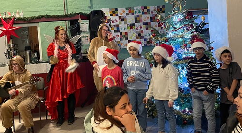 Weihnachtlich geschmückter Raum. 5 Kinder mit stehen vor einem festlichem Weihnachtsbaum. Eine Person im Renntierkostüm sitzt mit einer Gitarre in den Händen auf einem Stuhl. Ein Frau im roten Feenkostüm strahlt und klatscht. Alle sind sehr fröhlich.