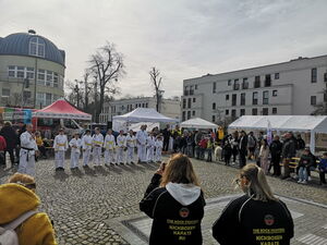 Großer Platz. Außen steh Gäste und in der Mitte eine Reihe Karateschüler in weißer Kleidung mit gelben bzw. weißen Gürtel.