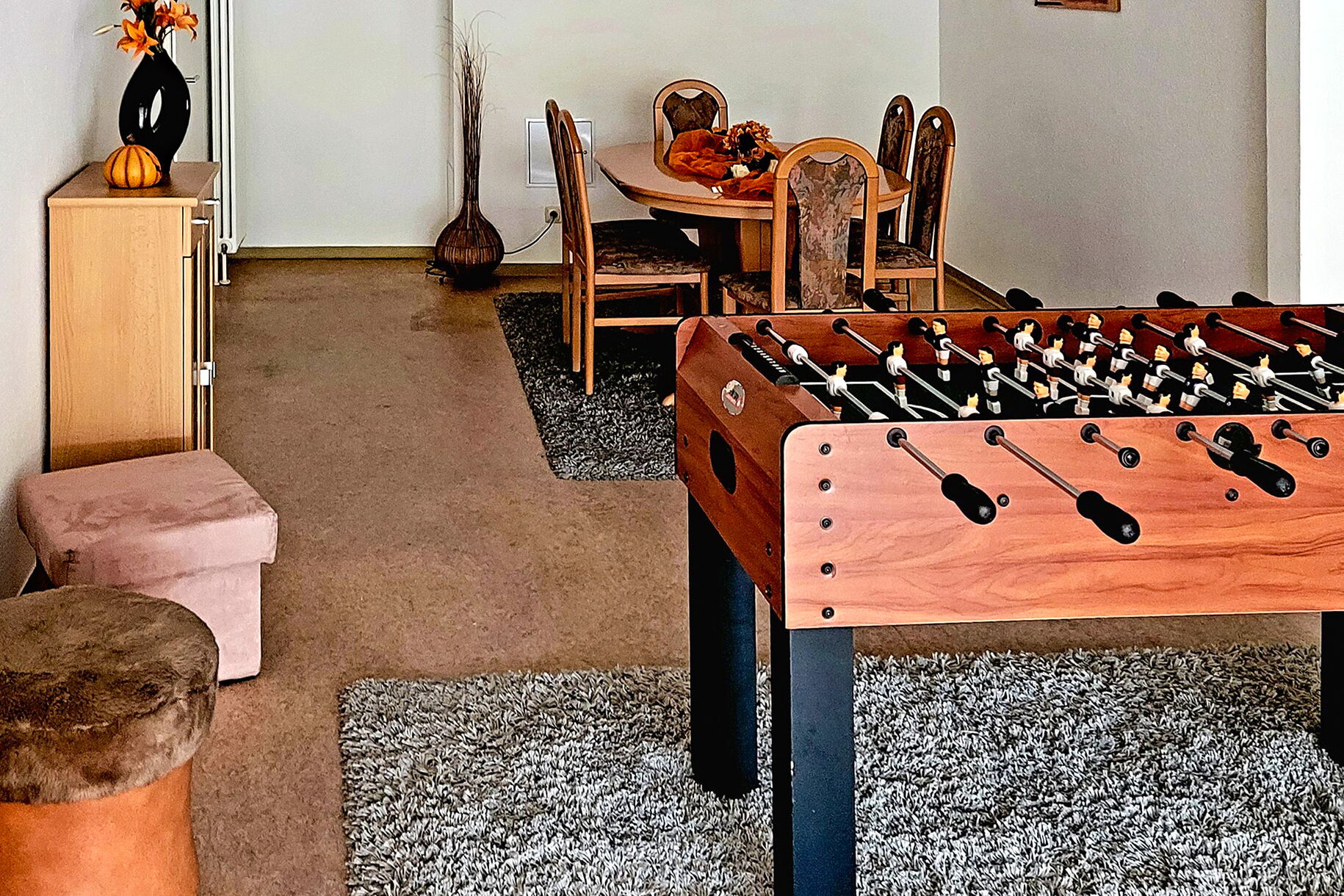 Tischkicker aus dunklen Holz auf grauem Teppich. Im Hintergrund ein ovaler Tisch mit 6 Stühlen steht auf auf einem grauen Teppich.