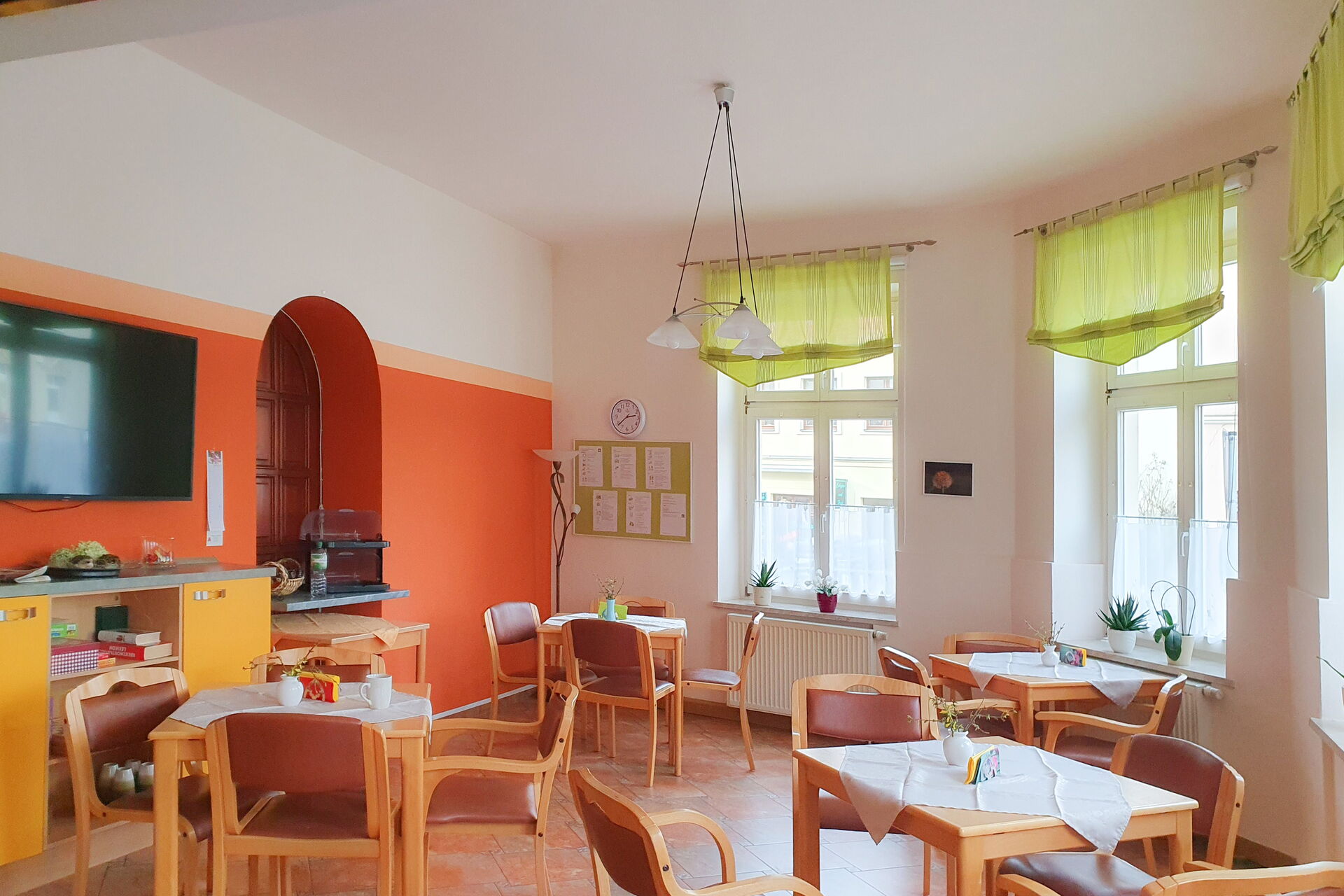 Speisesaal. Halbrunder Raum mit vier Tischen mit jeweils vier Stühlen. Heller freundlicher Raum, links: ein kleines Regal darüber ein Fernseher an einer Wand die orange und oben weiß ist.