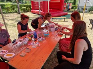 Draußen. Tisch an dem 4 Frauen sitzen und basteln. Die Sonne scheint. Eine rote Hüpfburg steht auf der Wiese.