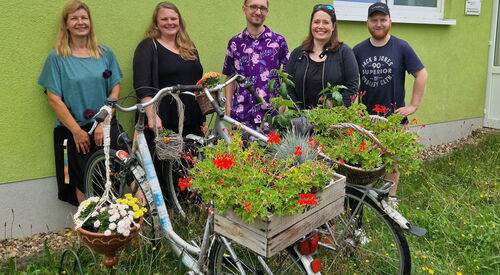 3 Frauen und zwei Männer stehen vor einer grünen Hauswand. Vor ihnen stehen zwei Fahrräder, auf den Gepäckträgern ist eine bepflanzte Holzkiste und ein bepflanter Korb mit Blumen. An einen Lenker hängt auch noch ein Körbchen mit Blumen.